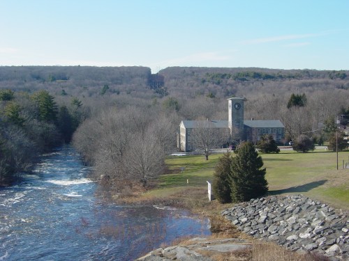 A serene landscape featuring a river, a clock tower building, and bare trees against a clear blue sky.