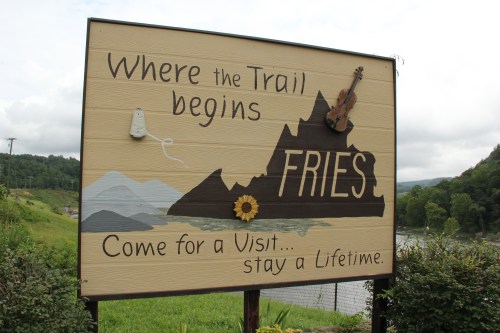 Sign reading "Where the Trail begins FRIES" with a mountain, violin, and sunflower, inviting visitors to stay a lifetime.
