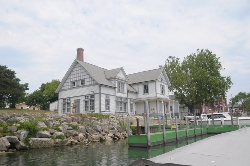 A large white house with a gray roof sits by the water, surrounded by greenery and rocky shoreline.