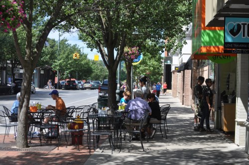 Outdoor café scene with people sitting at tables, trees lining the sidewalk, and shops in the background.