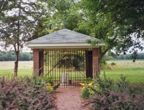 A small gazebo with a gated entrance, surrounded by greenery and flowers, set in a grassy field.