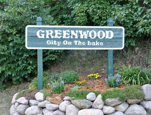 Sign for Greenwood, "City on the Lake," surrounded by flowers and rocks, set against a green backdrop.
