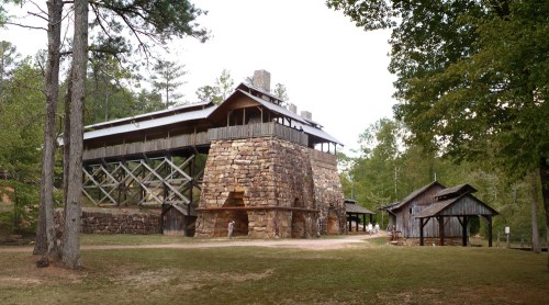 Historic stone structure with wooden upper level, surrounded by trees and additional rustic buildings.