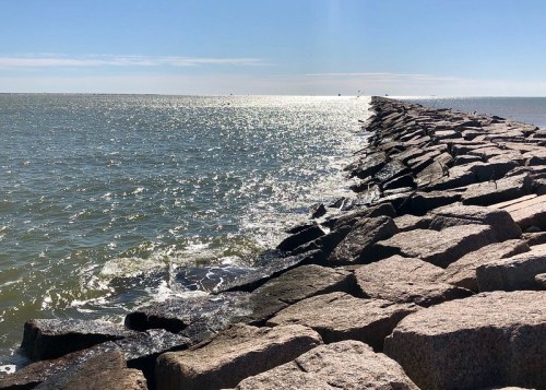 A rocky jetty extends into a shimmering sea under a clear blue sky, with gentle waves lapping at the stones.