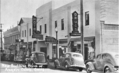 Black and white photo of a street in Angels Camp, California, featuring hotels, a drugstore, and vintage cars.