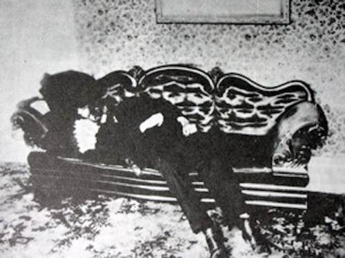 A black and white photo of two people reclining on a vintage couch in a patterned room.