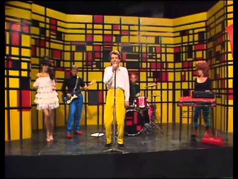 A colorful band performs on stage with a vibrant geometric backdrop, featuring a singer, guitarist, drummer, and keyboardist.