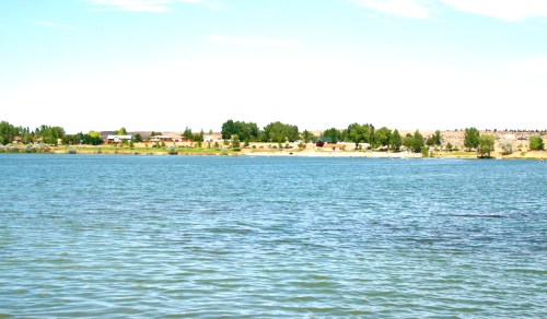 A calm lake with clear blue water, surrounded by green trees and a distant shoreline of buildings and grass.