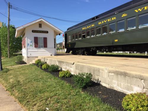 A vintage train passes by a small, white train station labeled "Flemington," surrounded by green grass and shrubs.
