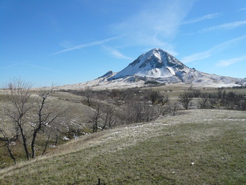 A snow-capped mountain rises above grassy hills under a clear blue sky, with scattered trees in the foreground.