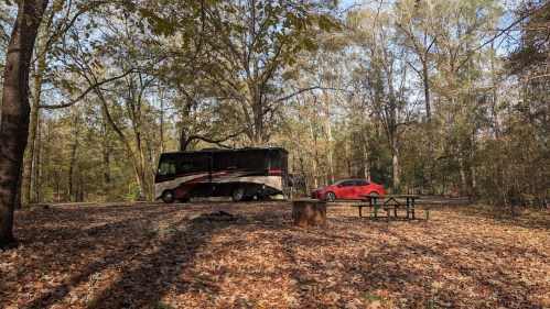 An RV parked in a wooded area with autumn leaves, next to a red car and a picnic table.