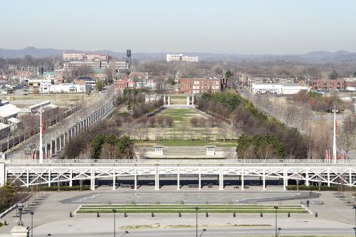 Aerial view of a park with pathways, greenery, and buildings in the background under a clear sky.