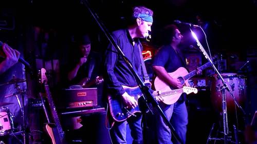 A live band performs on stage with guitars, drums, and keyboards in a dimly lit venue.