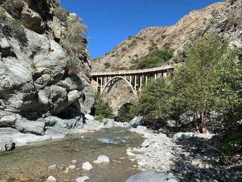 A stone bridge arches over a rocky riverbed, surrounded by steep hills and greenery under a clear blue sky.