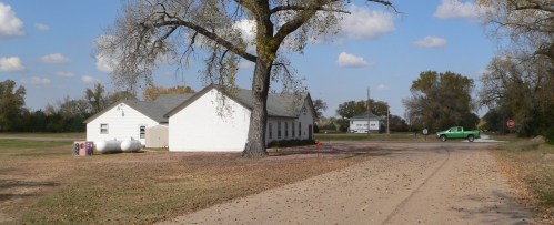 A rural scene featuring a white building, a large tree, and a green truck on a dirt road under a blue sky with clouds.