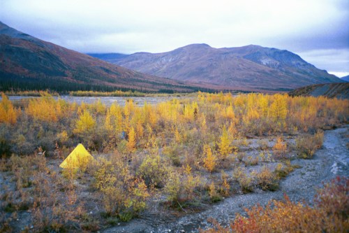 A yellow tent in a valley surrounded by autumn foliage and mountains under a cloudy sky.