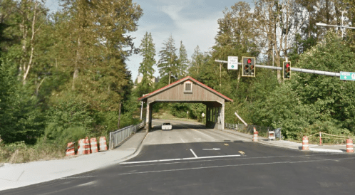 A covered bridge with traffic lights, surrounded by trees and construction barriers, leading to a road.