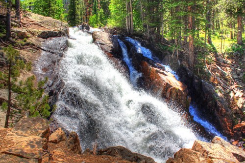A cascading waterfall flows over rocky terrain, surrounded by lush green trees in a serene forest setting.