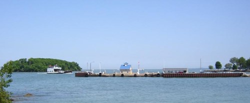 A calm waterfront scene featuring a pier, a small boat, and lush greenery in the background under a clear blue sky.