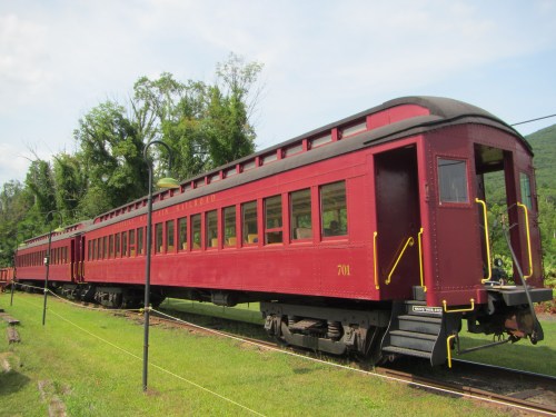 A vintage red train car parked on a grassy area, surrounded by trees and under a clear blue sky.