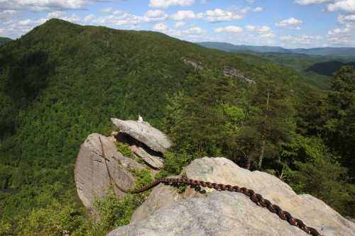 A rocky cliff with a chain leading down, surrounded by lush green mountains and a blue sky with fluffy clouds.