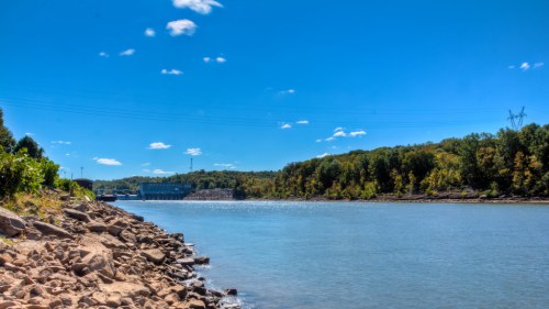 A serene river scene with rocky banks, lush trees, and a clear blue sky, featuring a distant dam and power lines.