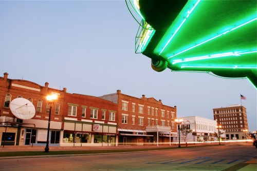 A quiet street scene at dusk featuring vintage buildings and a neon sign, with a hint of an American flag in the background.