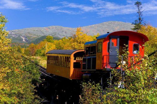 A colorful train with a red engine and yellow passenger car travels through a scenic landscape of autumn foliage and mountains.