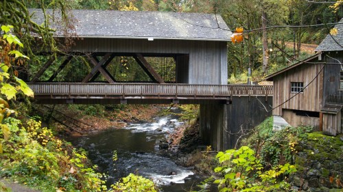 A wooden covered bridge spans a river, surrounded by lush greenery and autumn foliage.