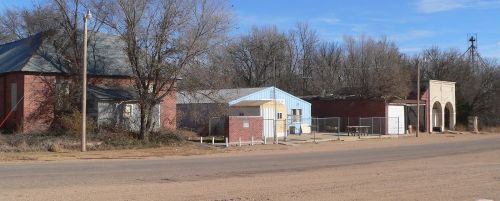 A quiet rural scene featuring a few buildings along a dirt road, surrounded by trees under a clear blue sky.