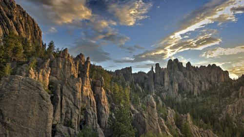 A scenic view of rugged rock formations and pine trees under a colorful sky at sunset.