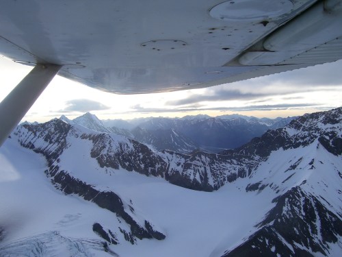 Aerial view of snow-covered mountains and glaciers under a cloudy sky, seen from an airplane wing.