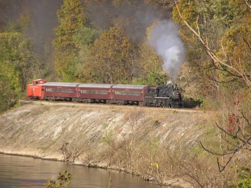 A vintage steam train with a red caboose travels along a riverbank surrounded by autumn trees.