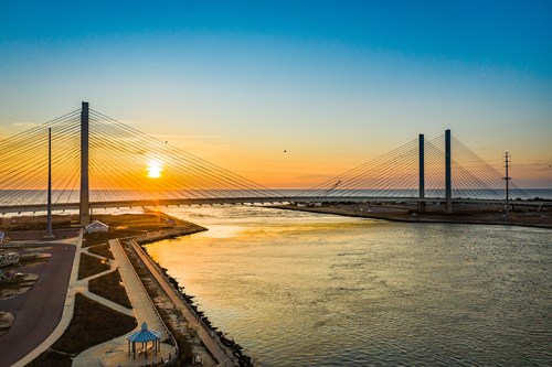 A sunset over a bridge with cable supports, reflecting on a calm river, surrounded by a scenic landscape.