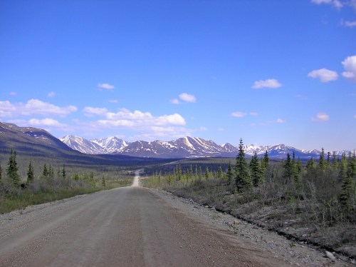 A dirt road stretches through a mountainous landscape under a blue sky with scattered clouds. Snow-capped peaks in the distance.
