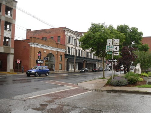 A rainy street scene featuring brick buildings, street signs, and parked cars in a small town.