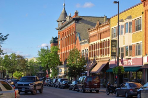 A charming downtown street lined with historic buildings, trees, and parked cars under a clear blue sky.
