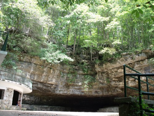 A rocky cave entrance surrounded by lush green trees and a stone structure in the foreground.