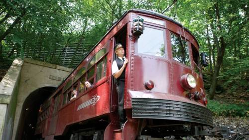 A vintage red train enters a tunnel, with a conductor at the front and passengers visible in the windows.