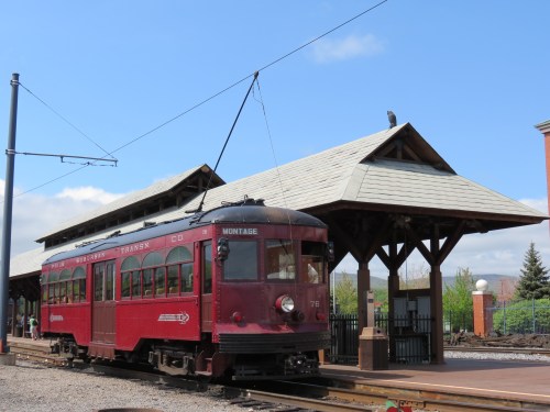 A vintage red tram at a train station with a wooden shelter and blue sky in the background.