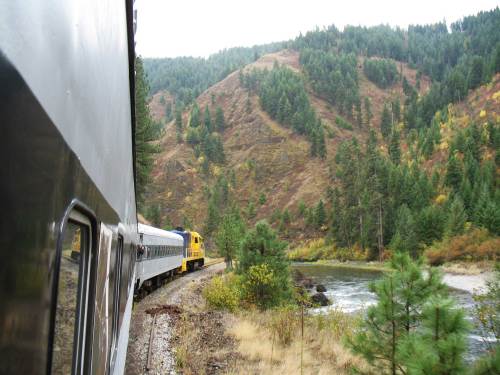 A train winds along a riverbank, surrounded by lush green hills and autumn foliage.