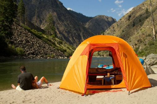 A bright orange tent by a river, with mountains in the background and two people relaxing on the sandy shore.