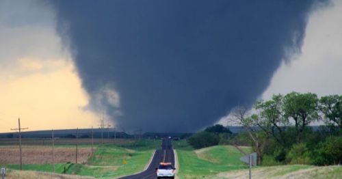 A large tornado looms over a rural road, with a vehicle parked nearby and dark storm clouds in the sky.
