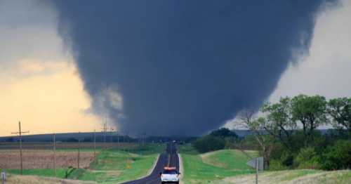 A large tornado looms over a rural road, with a vehicle parked nearby and dark storm clouds in the sky.