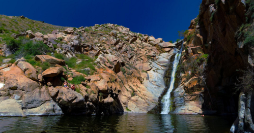 A serene waterfall cascades over rocky cliffs into a calm pool, surrounded by lush greenery and clear blue skies.
