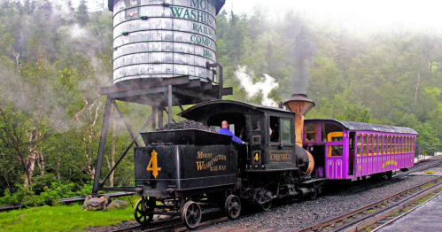 A vintage steam train with a black engine and purple passenger cars, passing by a water tower in a scenic landscape.