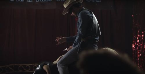 A cowboy in a hat performs on stage, with a spotlight highlighting the scene in a dimly lit venue.