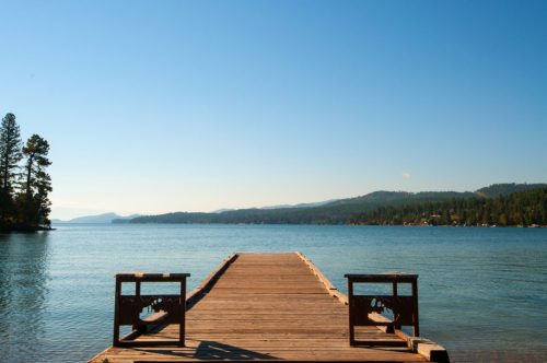 A wooden dock extends into a calm lake, surrounded by trees and mountains under a clear blue sky.