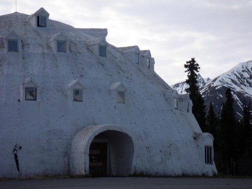 A large, dome-shaped white building with small windows, surrounded by trees and mountains under a cloudy sky.