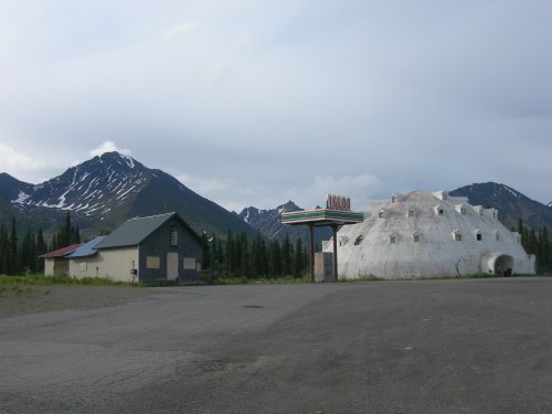 A unique white igloo-shaped building next to a rustic cabin, set against a backdrop of mountains and cloudy skies.